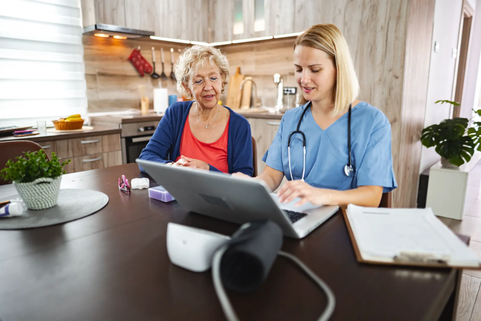 Healthcare professional with a stethoscope showing information on a laptop to an elderly woman in a kitchen.