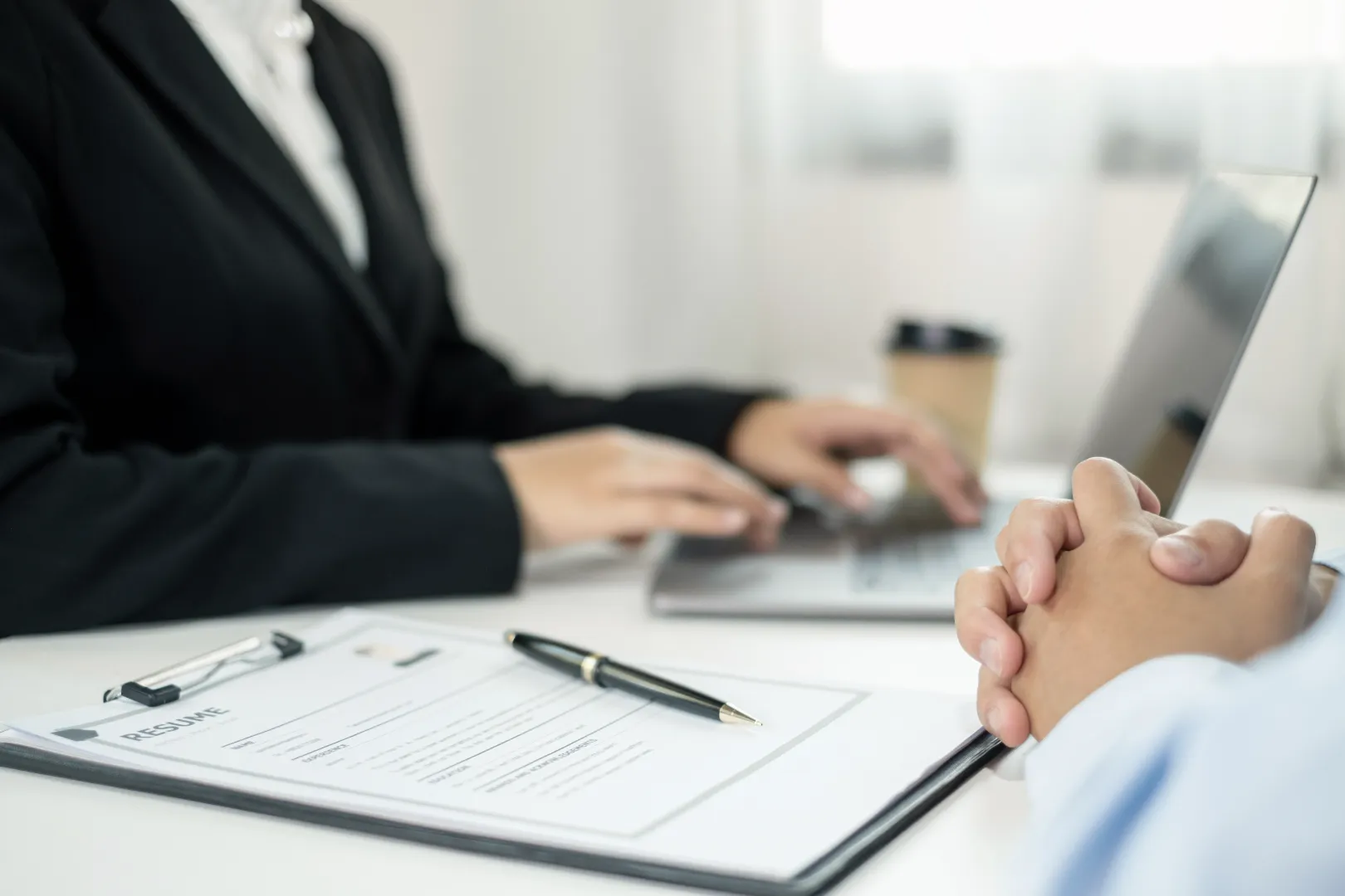Job interviewer typing on a laptop with a resume and pen on the table, and interviewee's hands clasped in the foreground.