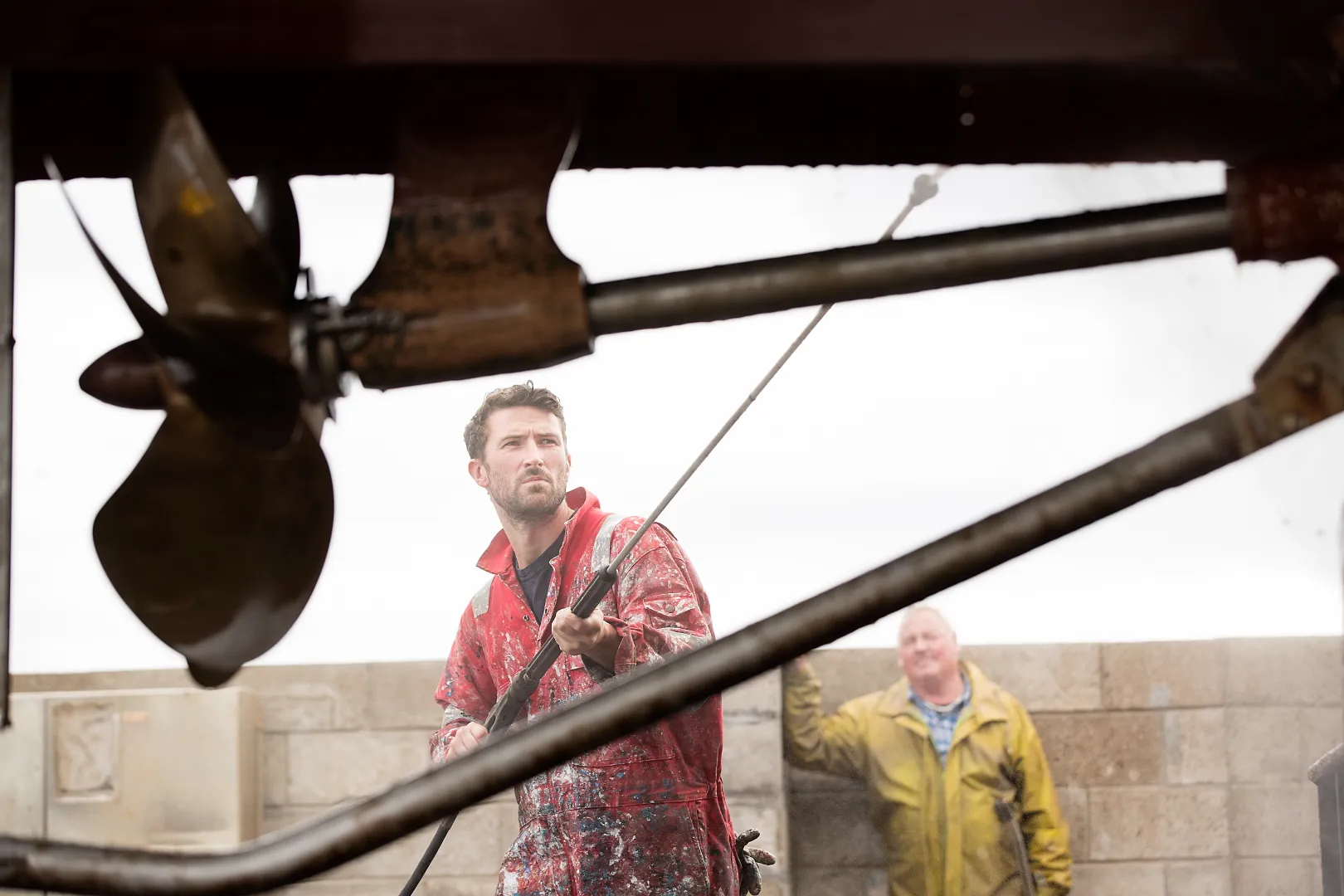 Man in red protective coverall pressure washing a boat propeller with another man in a yellow raincoat in the background.