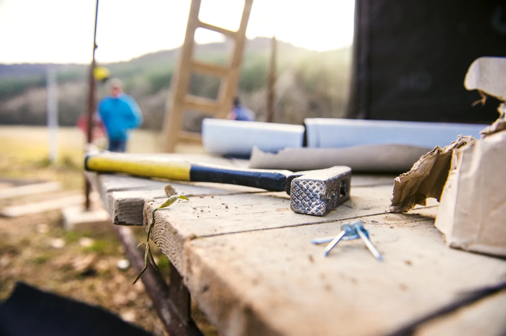 Hammer with yellow and black handle lying on a wooden plank table at a construction site, with nails and cardboard nearby.