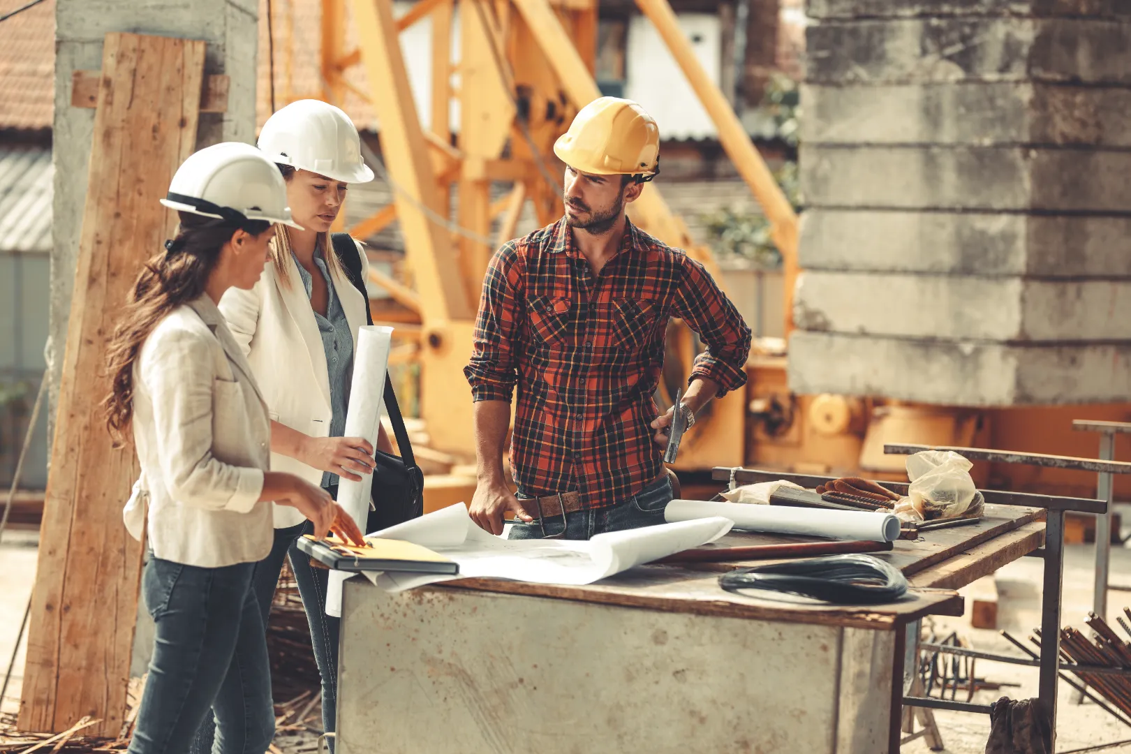 Three construction professionals wearing hard hats discussing blueprints on a table at a construction site.