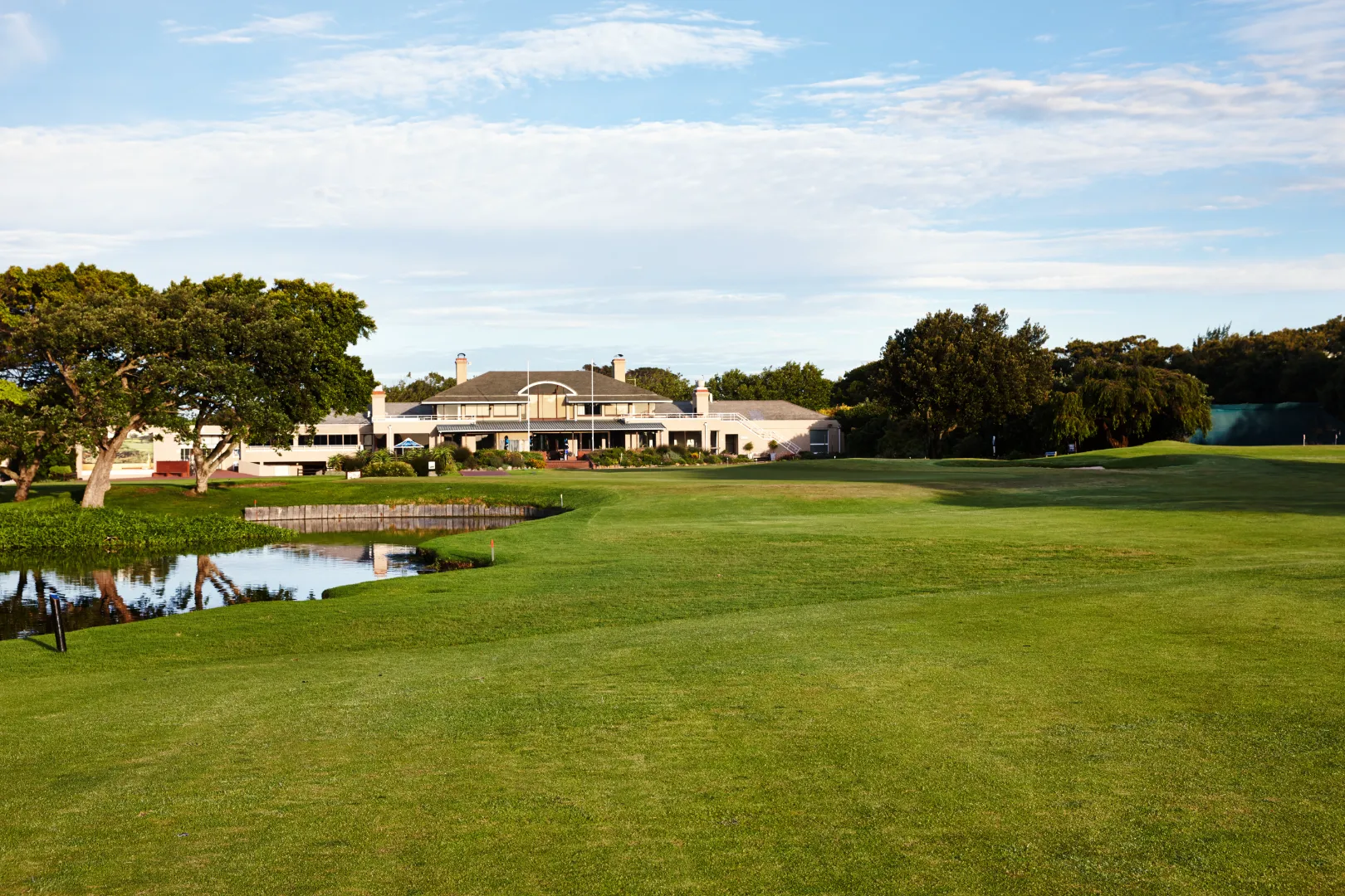 Golf course with green fairway, water hazard on the left, large clubhouse in the background, and trees under a partly cloudy sky.