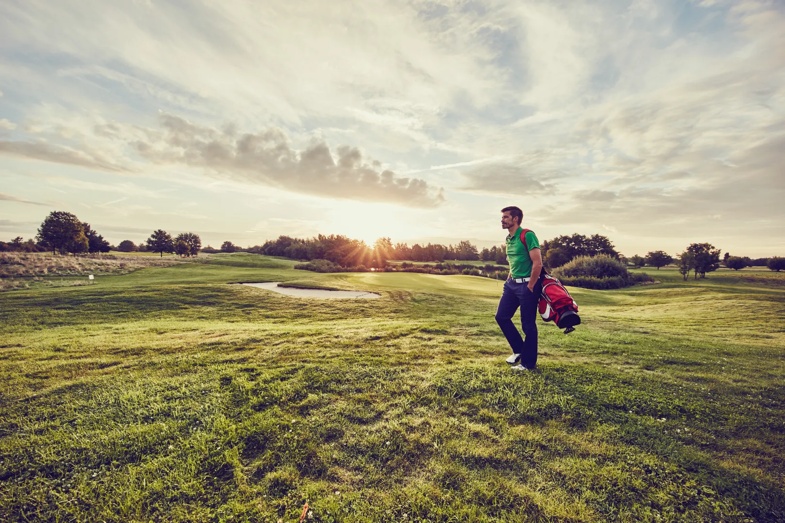 Man in green shirt walking on a golf course carrying a red golf bag at sunset.