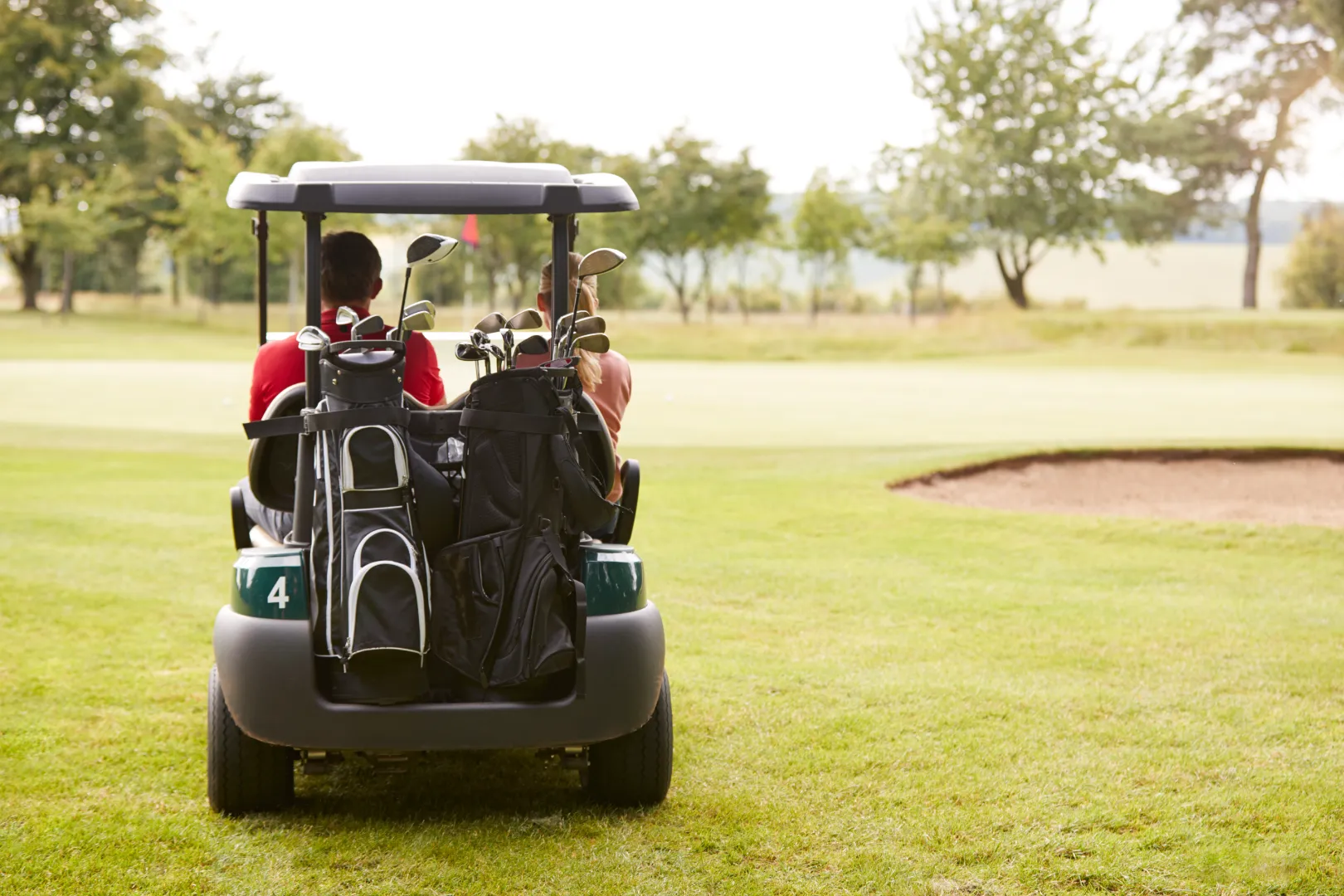 Two people sitting in a golf cart with golf bags on the back, facing a putting green with a sand bunker on the right.