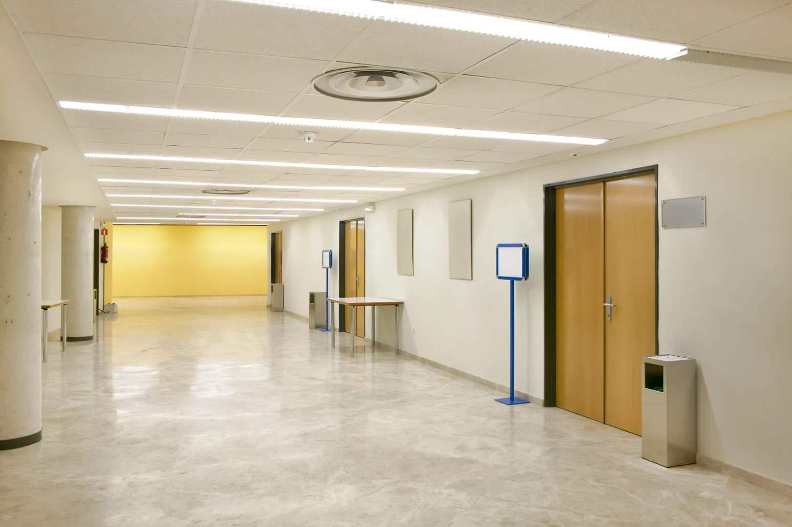 Long, empty hallway with polished floors, beige walls, wooden doors, blue sign stands, and fluorescent ceiling lights.