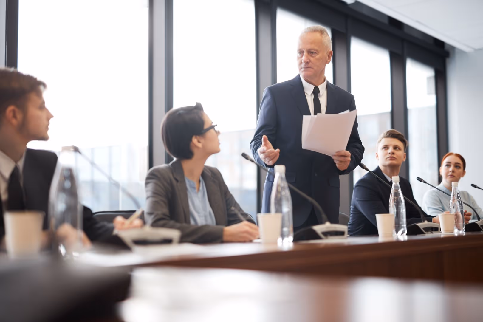 Man in suit standing and speaking while holding papers at a conference table with attentive colleagues seated around him.