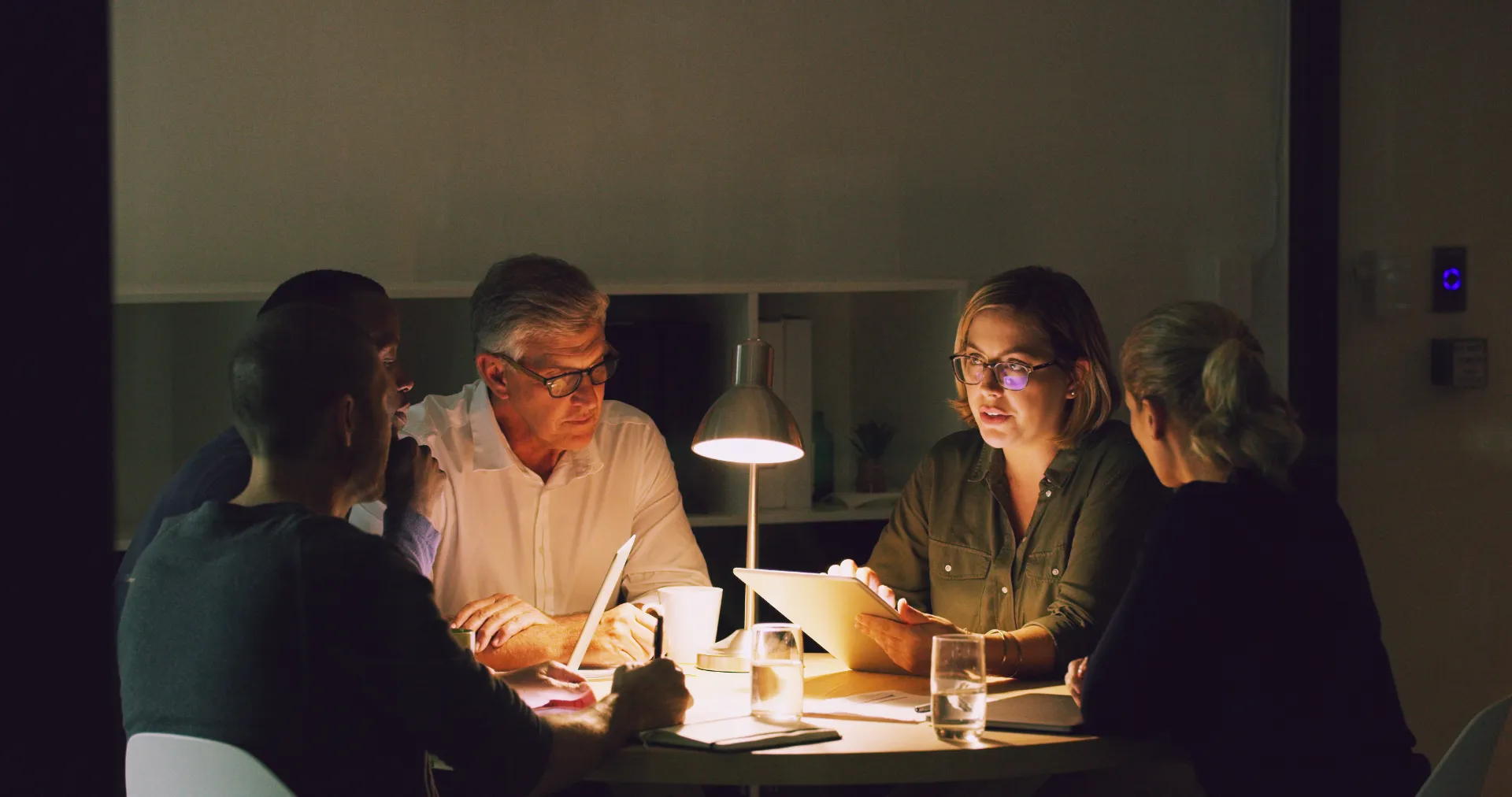 Five people having a serious discussion around a table illuminated by a desk lamp in a dimly lit room.