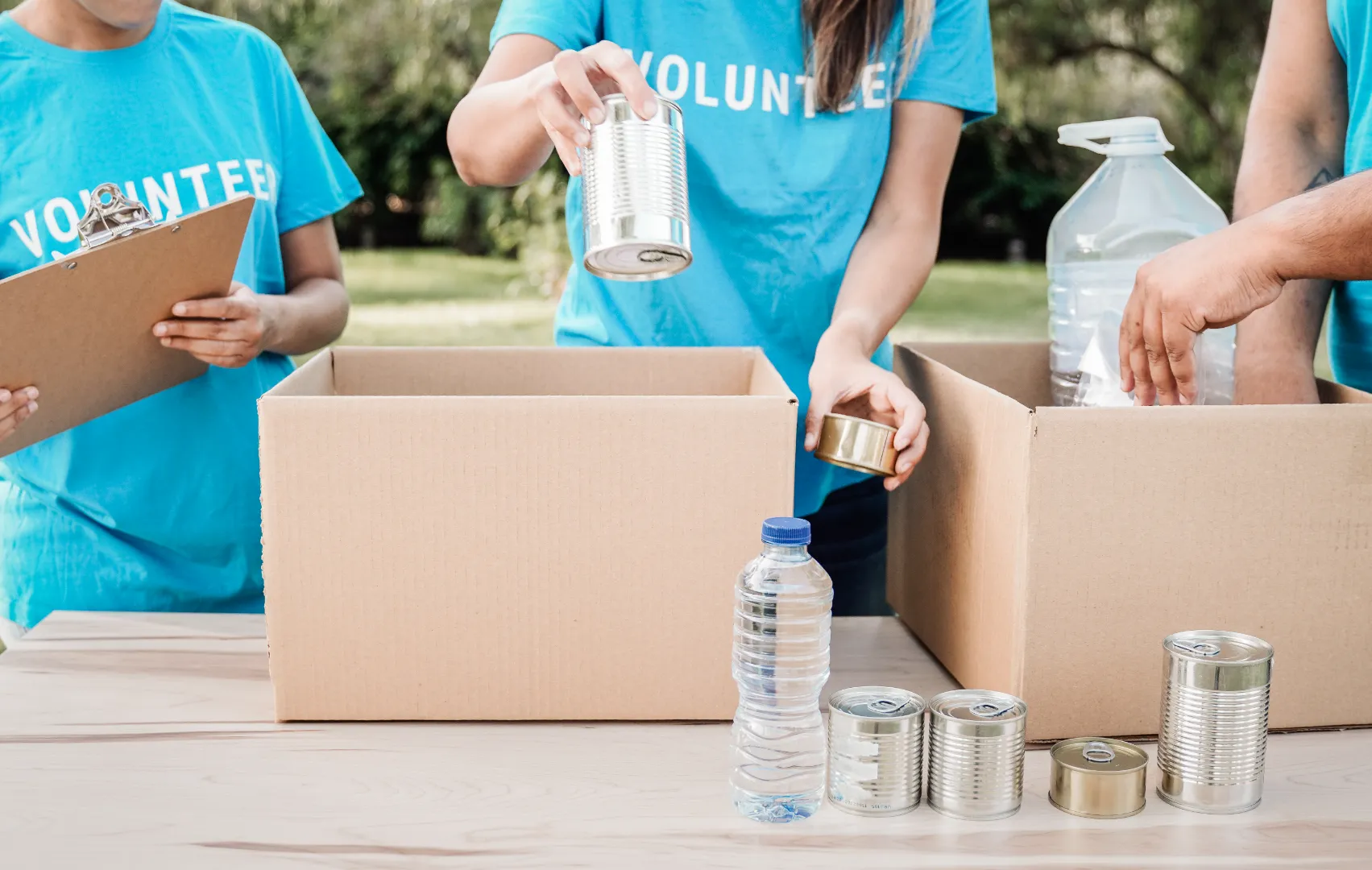 Volunteers in blue shirts packing canned food and bottled water into cardboard boxes outdoors.