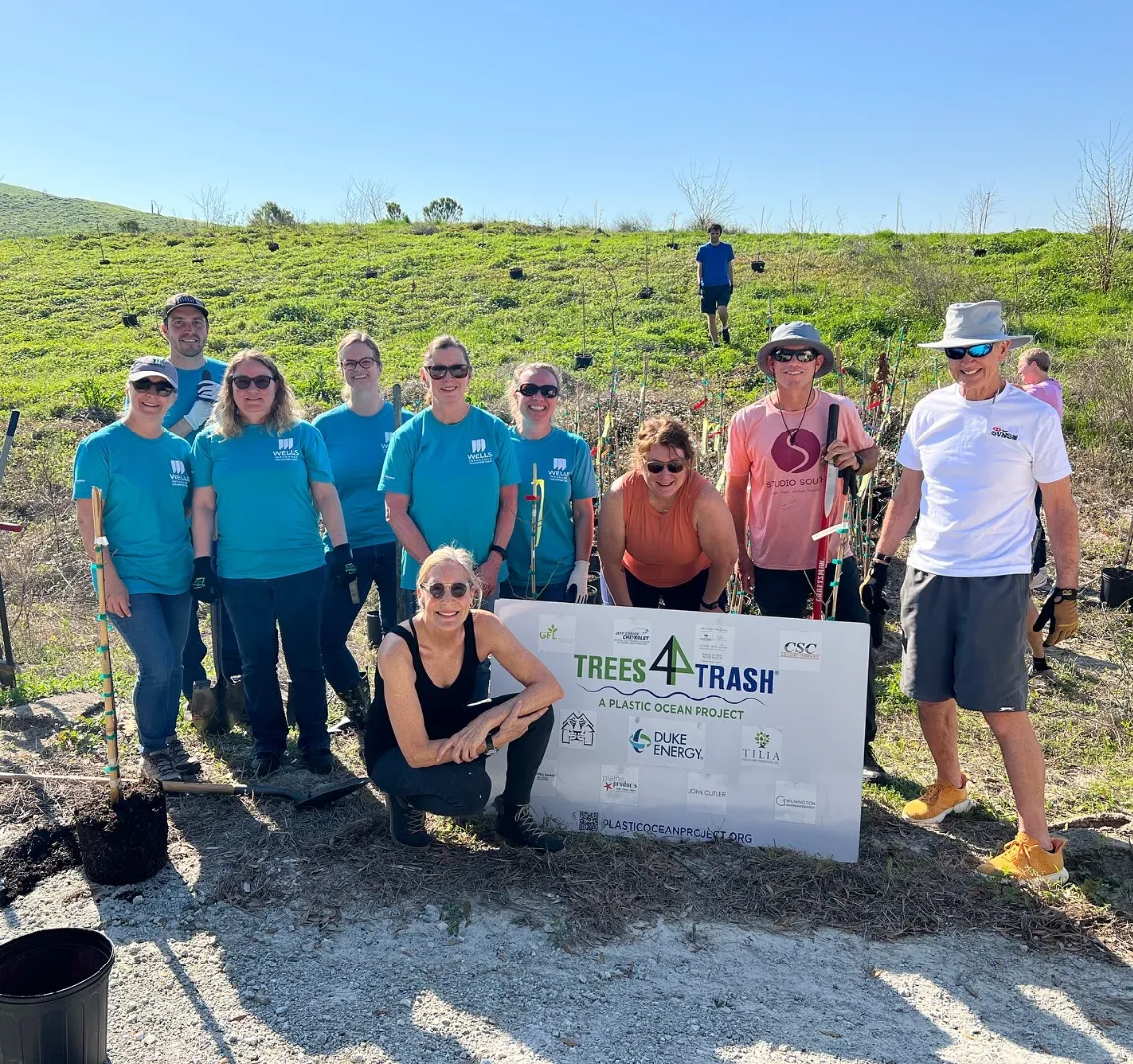 Group of volunteers posing outdoors on a sunny day with trees and planting tools behind a sign that reads 'Trees 4 Trash, A Plastic Ocean Project'.