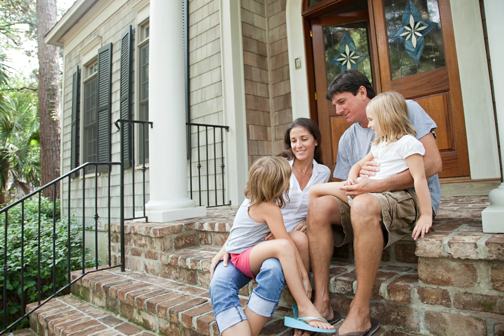 Family of four sitting on brick steps outside a house, smiling and interacting.