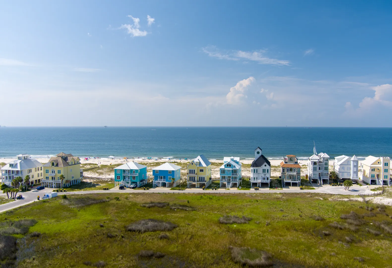 Colorful beachfront houses along a sandy beach with green grass in the foreground and a calm ocean under a blue sky.