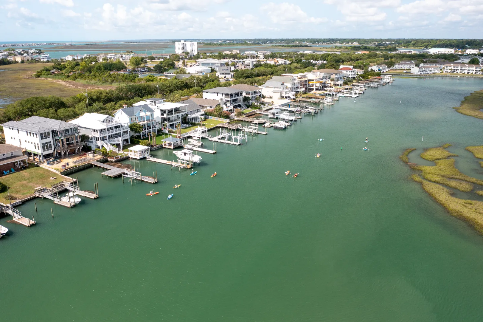 Aerial view of a coastal residential area with docks, boats, and kayakers paddling on calm water.
