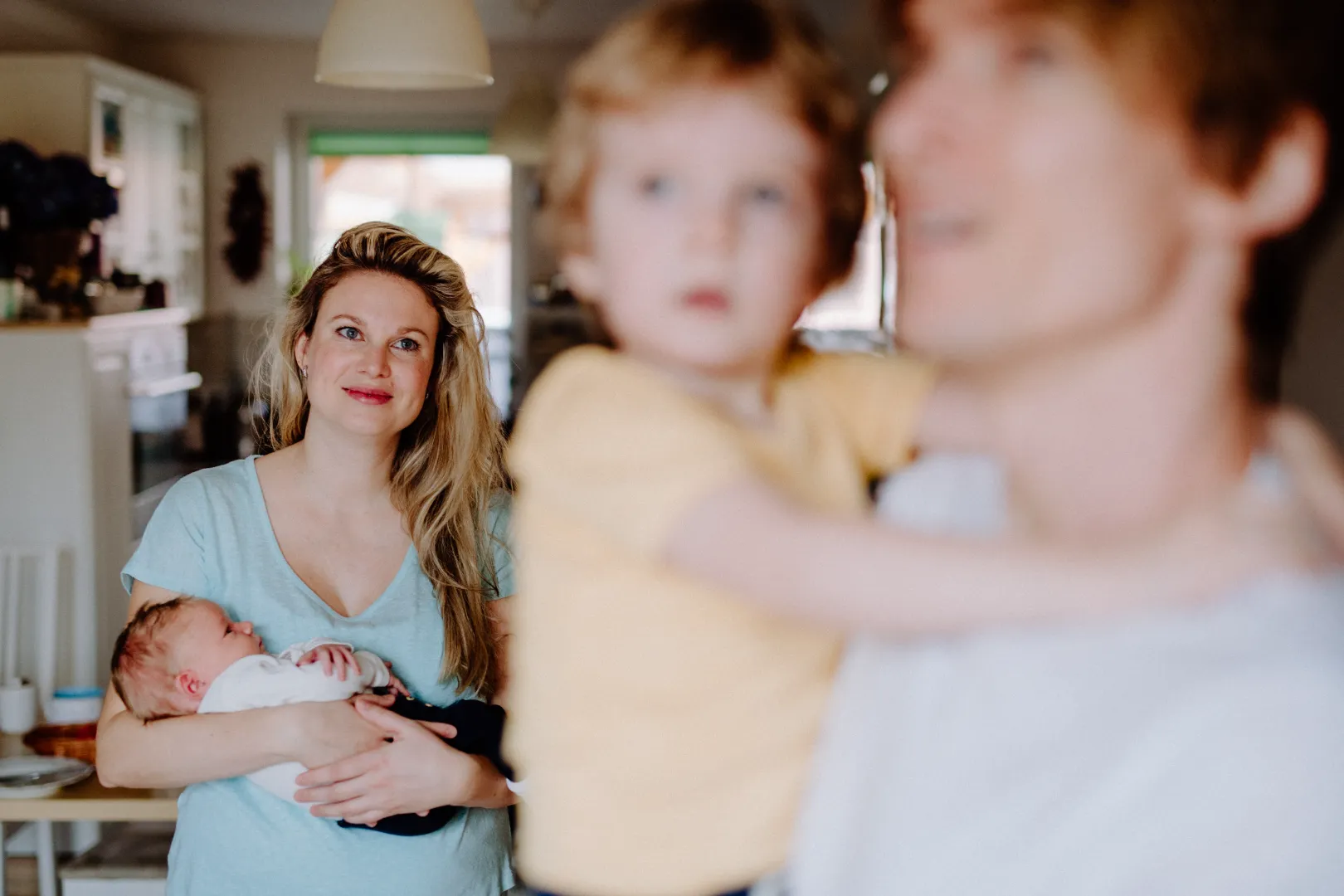 Smiling woman holding a sleeping newborn in a bright home kitchen, with a blurred man holding a child in the foreground.