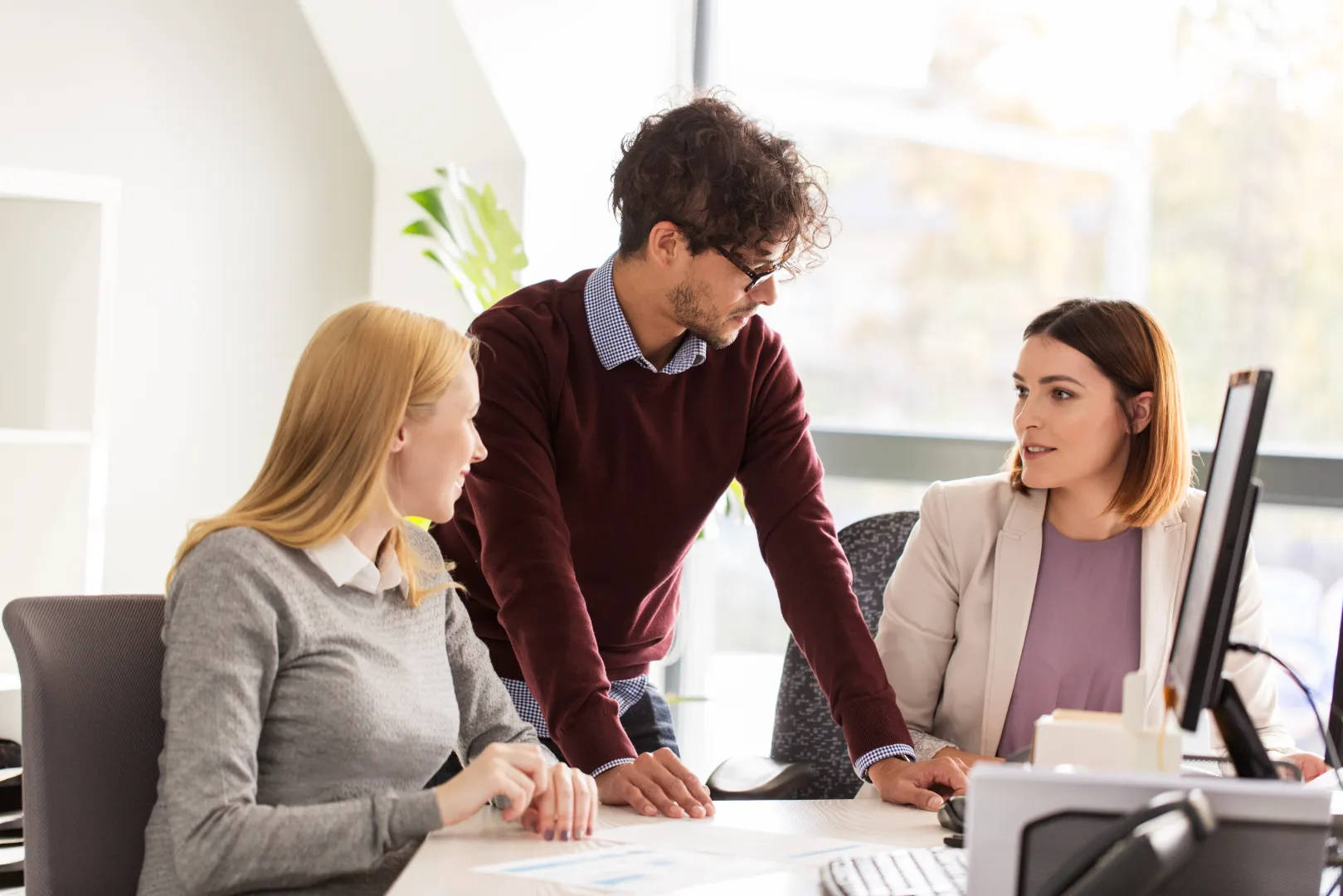 Three colleagues in an office discussing work, with one man standing and two women seated at a desk with a computer.