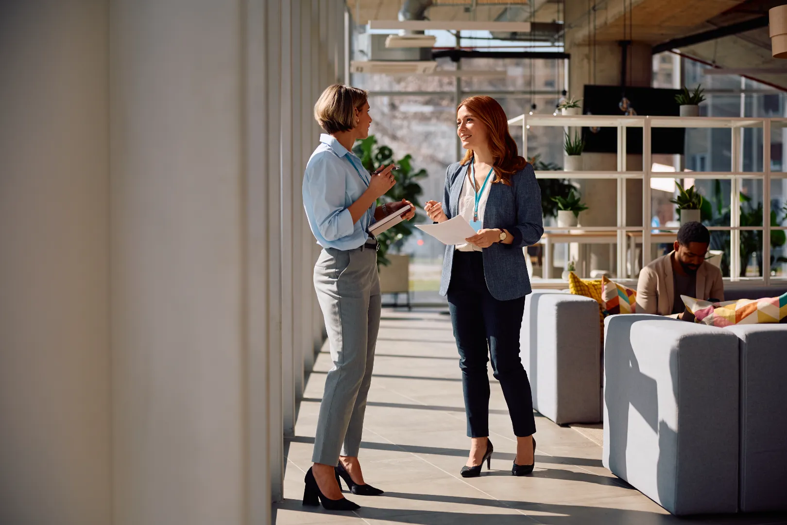Two professional women conversing in a bright modern office hallway, one holding a notebook and the other holding papers, while a man works on a laptop seated on a sofa in the background.