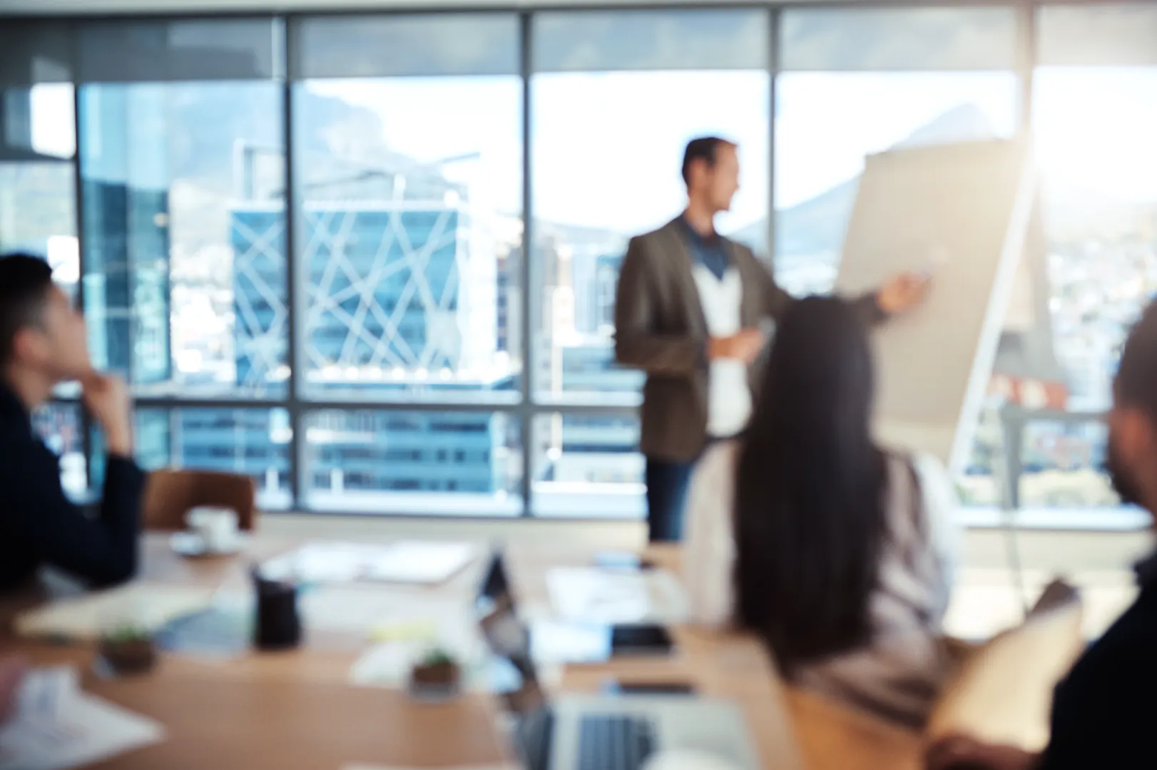 Blurry image of a business meeting with a presenter standing by a flip chart and attendees seated at a table in a bright office with large windows.