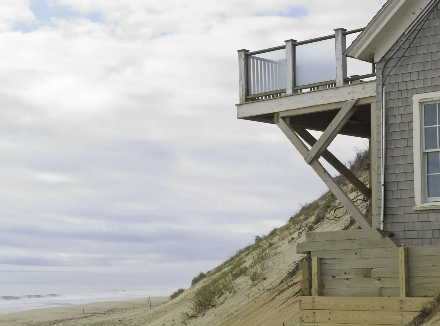 Wooden beach house supported by beams on a sand dune overlooking a cloudy shoreline.