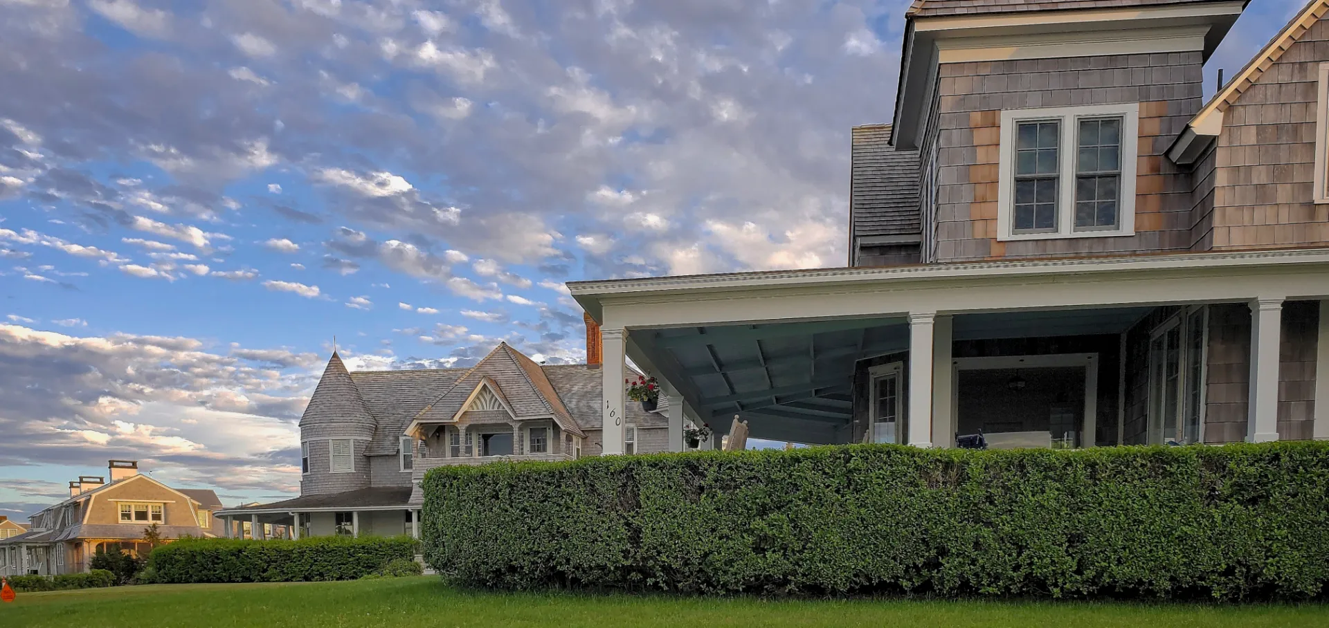 Large gray shingled houses with porches and manicured hedges under a partly cloudy sky.