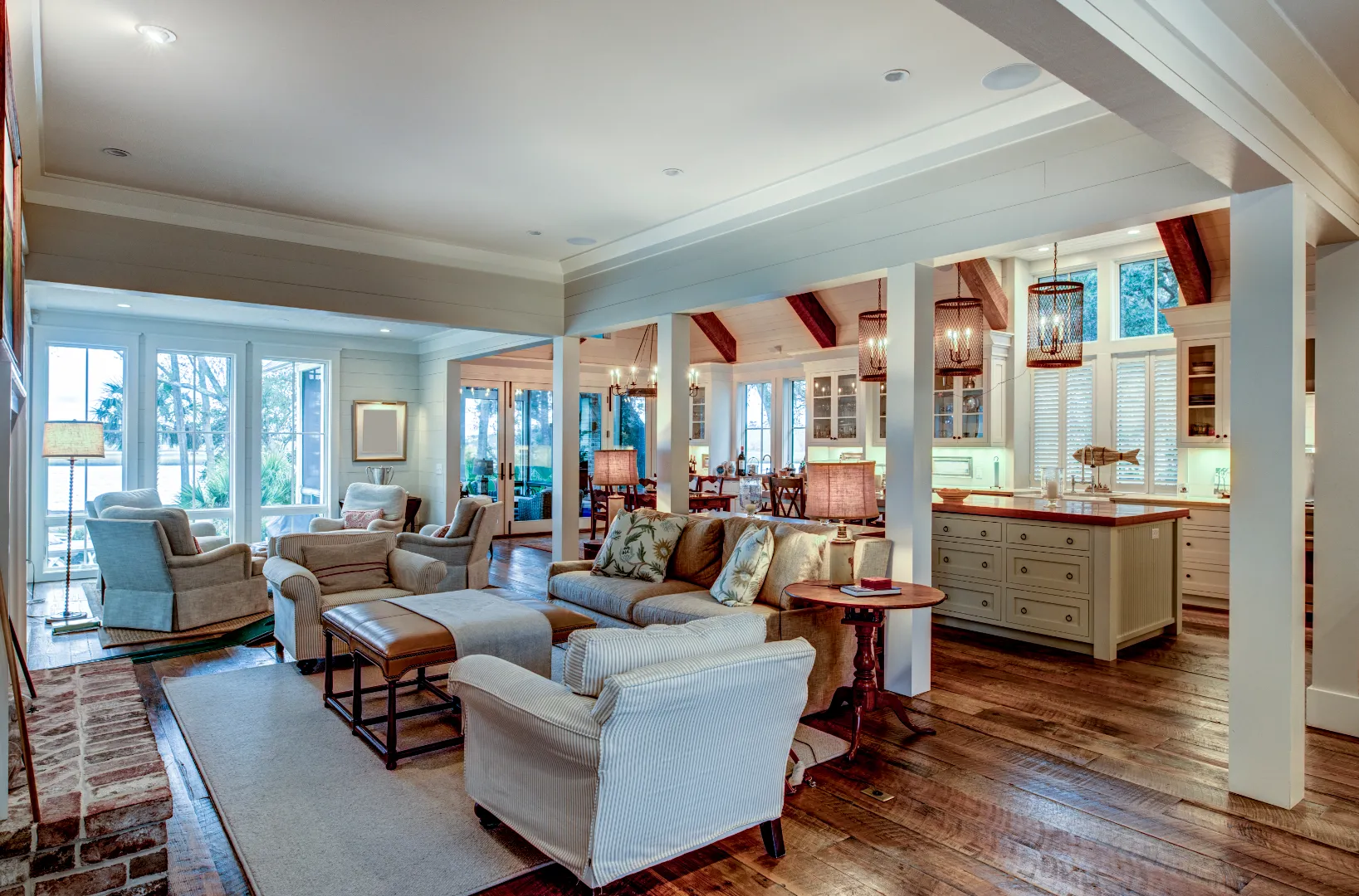 Open-concept living room with beige and striped armchairs, a sofa with floral pillows, ottoman, wooden floor, and adjoining kitchen with island and hanging pendant lights.