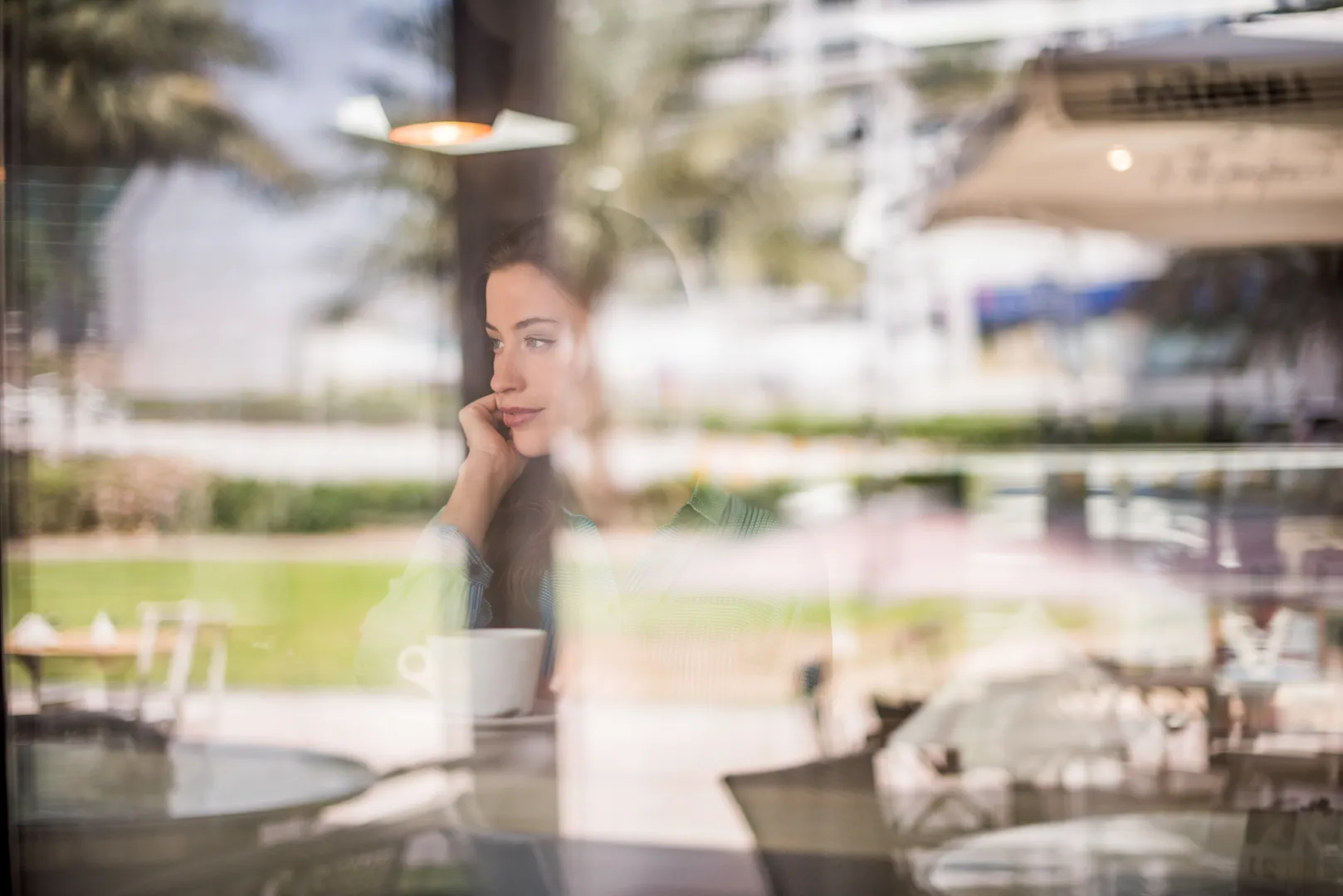 Woman sitting inside a cafe looking thoughtfully out the window with a cup of coffee.