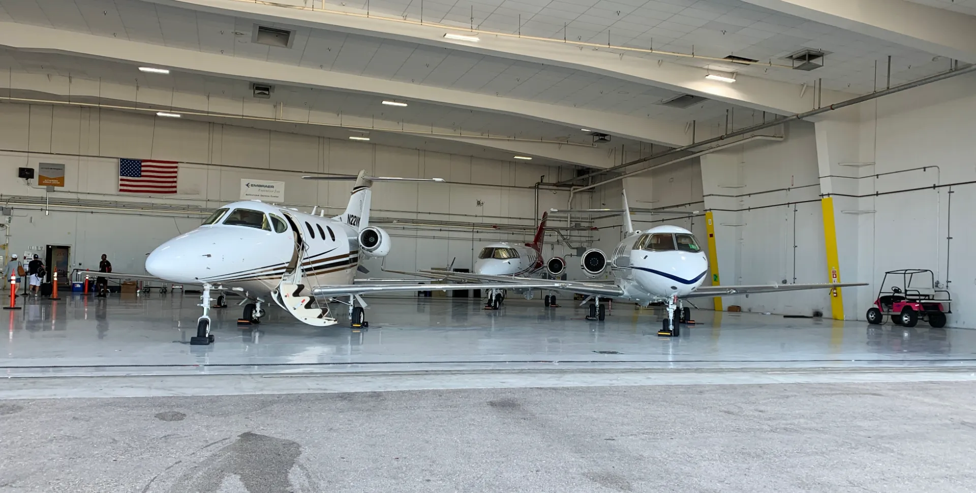 Three small private jets parked inside an aircraft hangar with an American flag on the wall and an open jet door on the left plane.