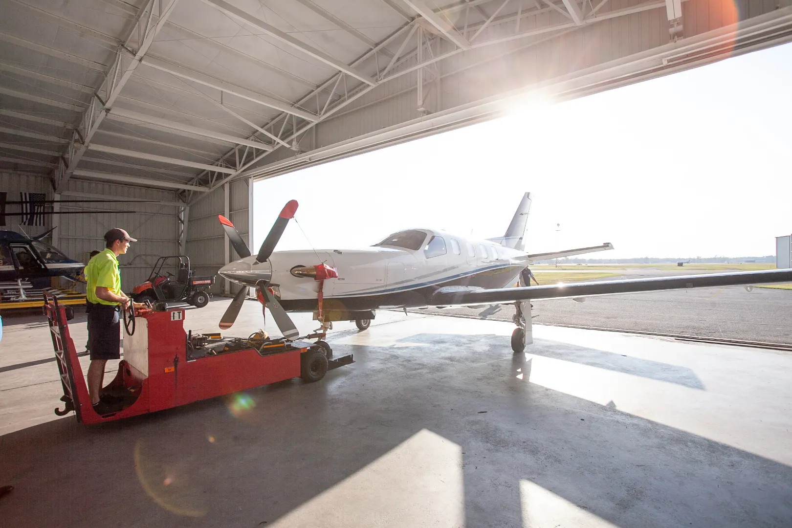 Man operating a red tug vehicle moving a small white propeller airplane out of a hangar on a sunny day.
