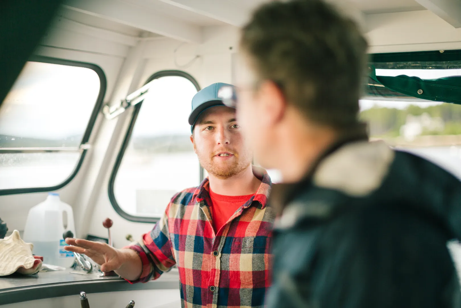 Two men inside a boat cabin, one wearing a blue cap and plaid shirt speaking to the other.
