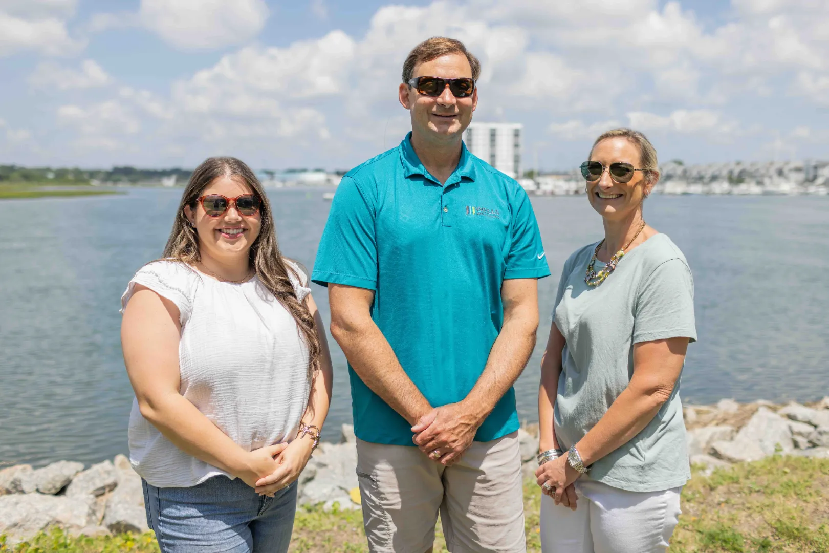Three smiling adults wearing sunglasses standing outdoors by a body of water on a sunny day.