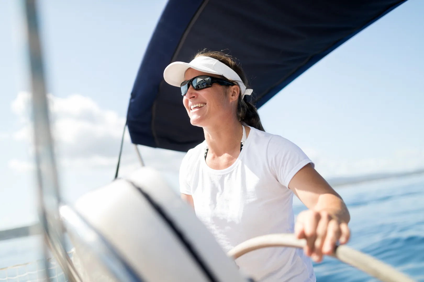 Smiling woman wearing sunglasses and a white visor steering a sailboat on a sunny day.