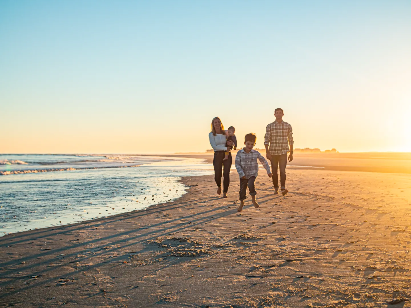 Family of four walking barefoot on a sandy beach at sunset with ocean waves in the background.