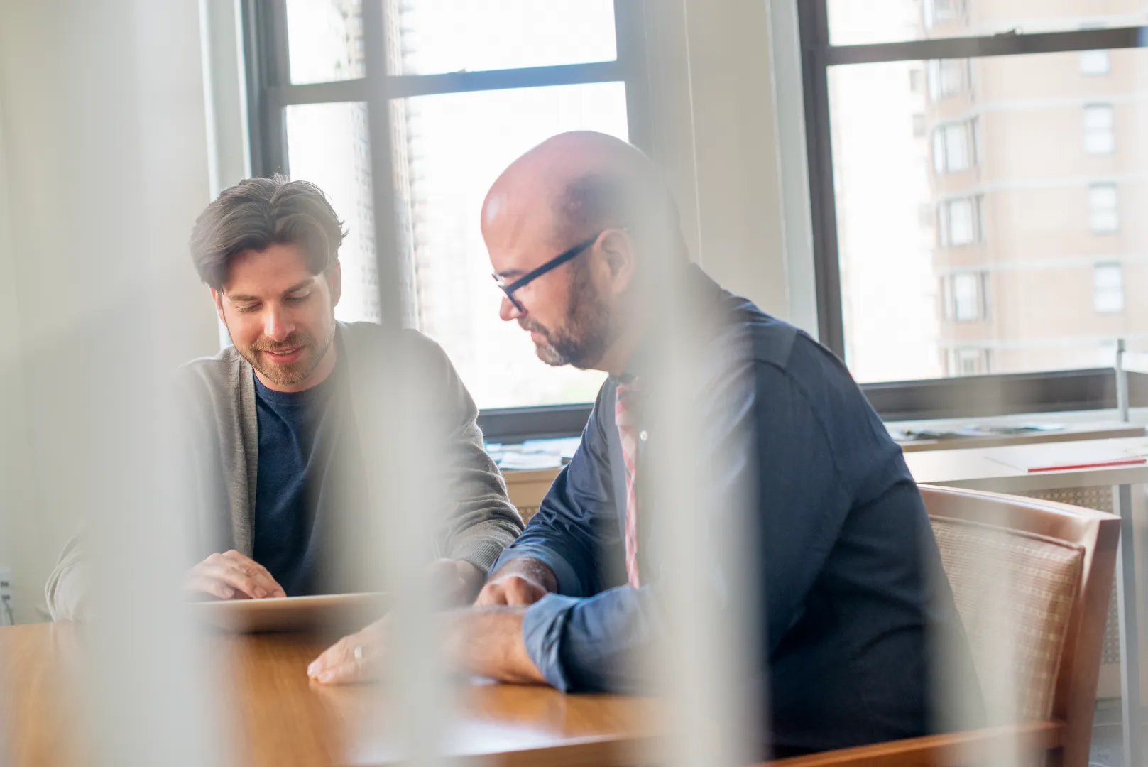 Two men sitting at a table in a bright office, looking at a tablet together.