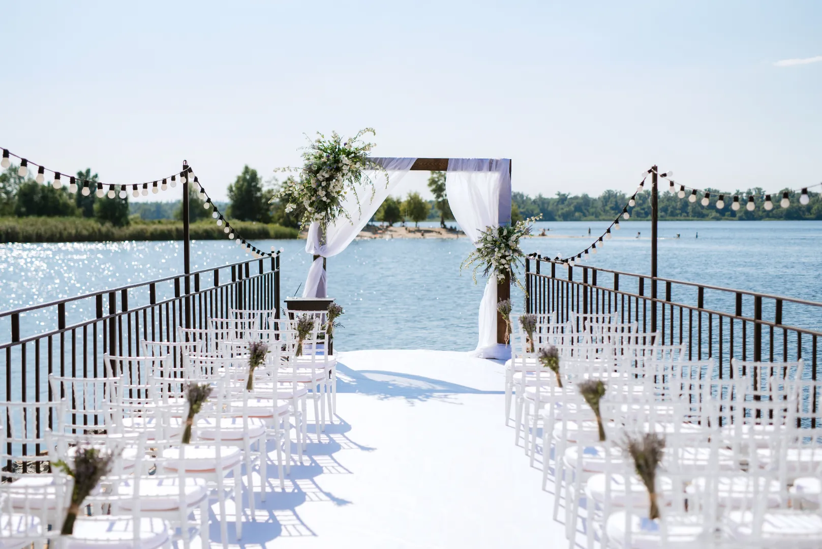 Outdoor wedding setup on a pier over water with white chairs, floral decorations, and a draped arch.