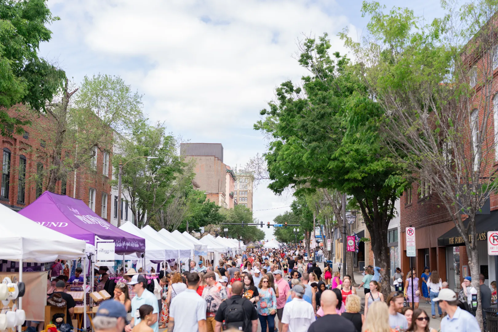 Crowded outdoor street market with white tents and people browsing under a partly cloudy sky.