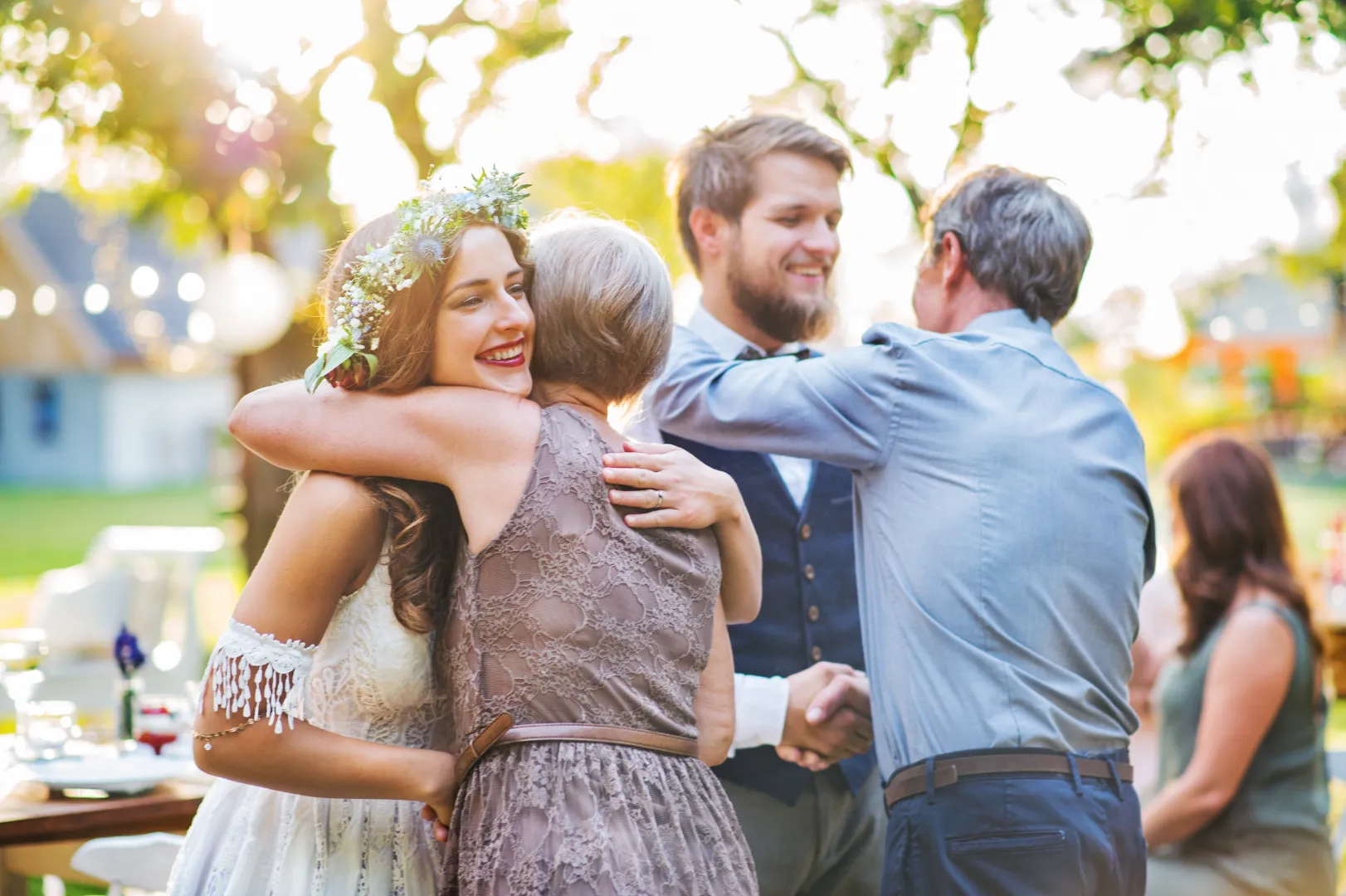 Bride with floral crown hugging an older woman while groom shakes hands with an older man at an outdoor wedding.