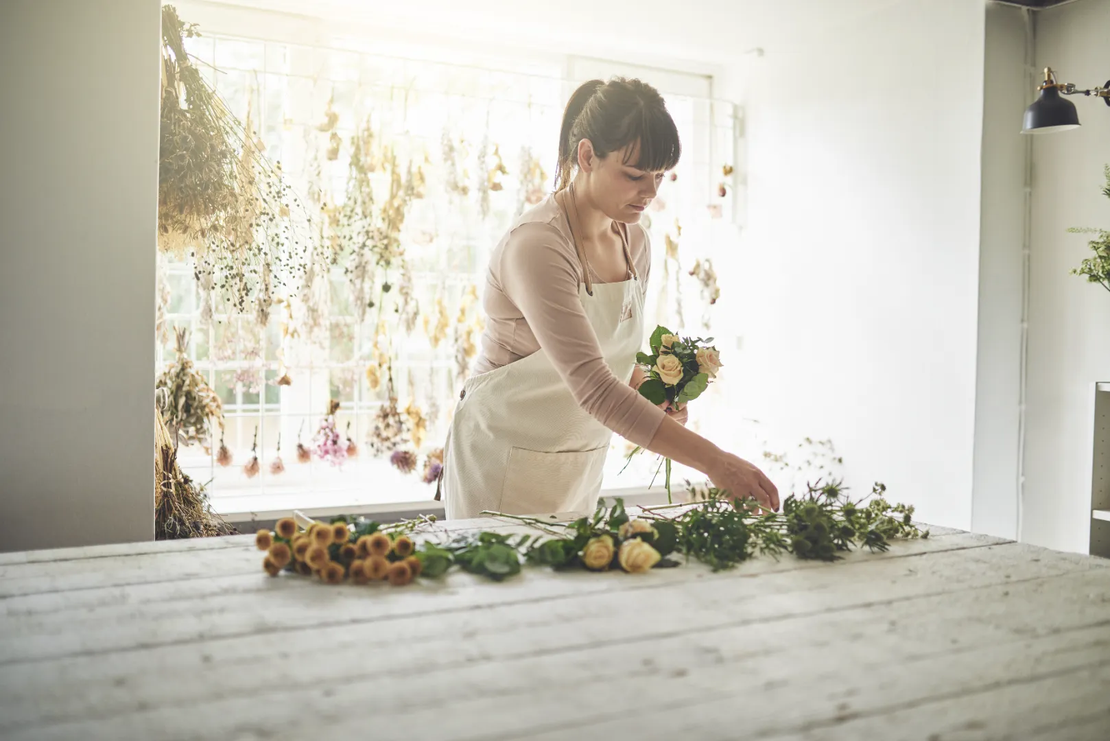 Woman arranging flowers on a table in a bright room with dried flowers hanging in the background.