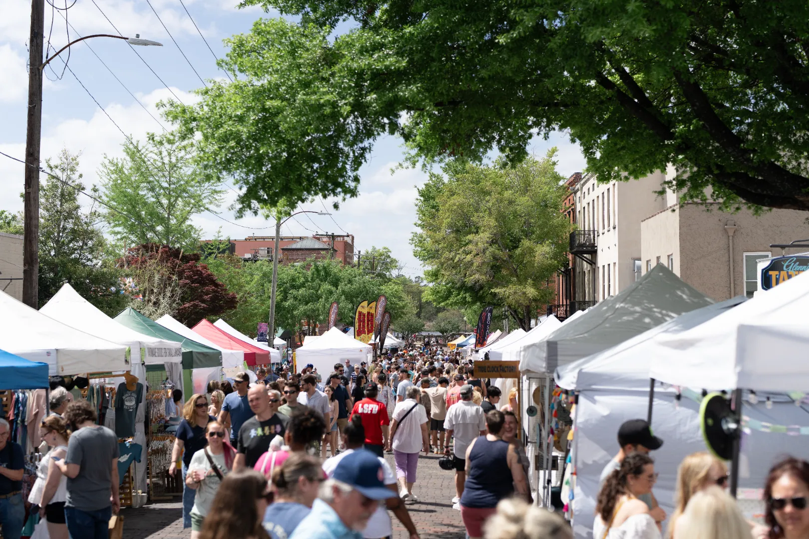 Busy outdoor street market with white tents and people walking under green trees on a sunny day.
