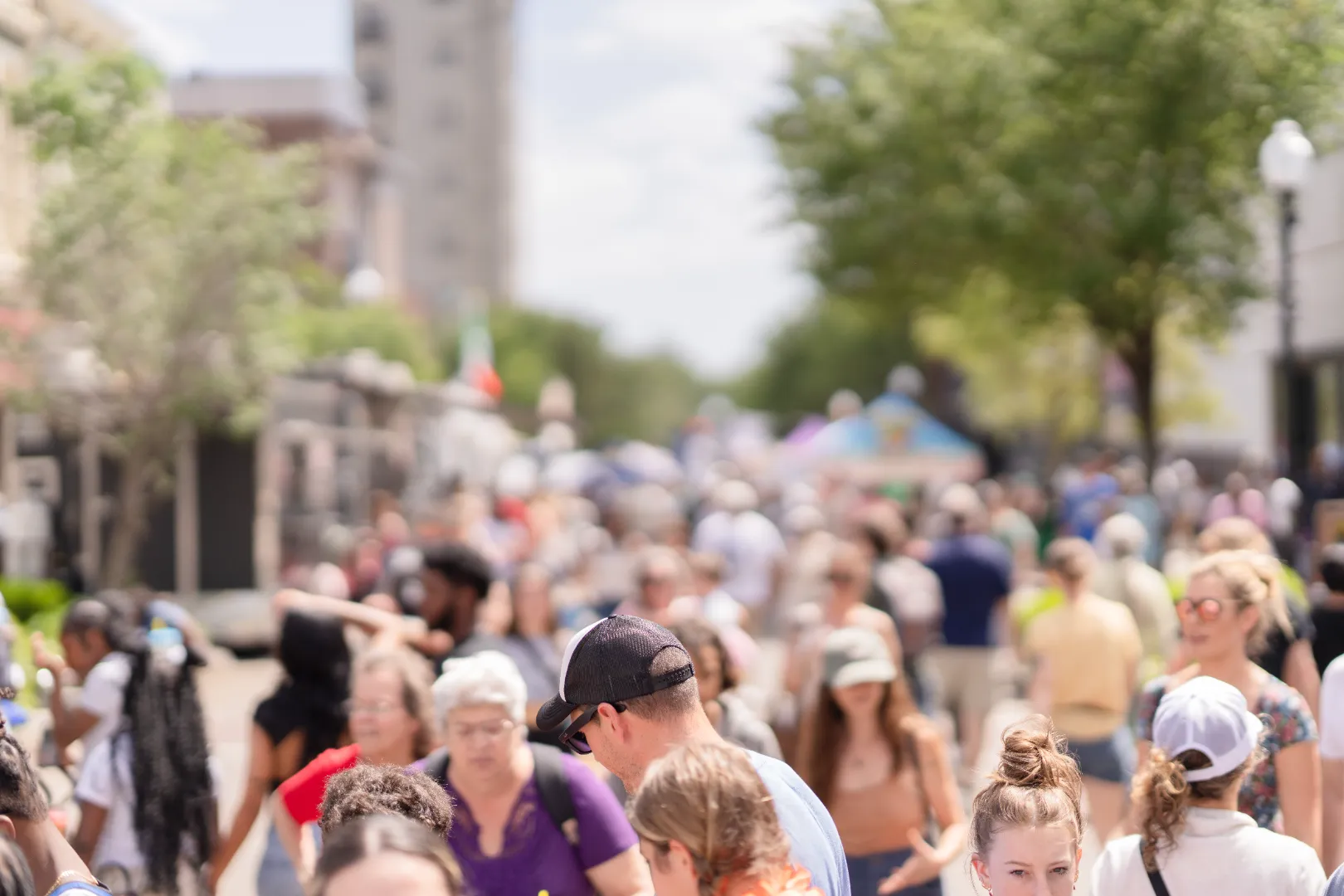 Crowd of people walking along a sunny street with trees and buildings in the background.