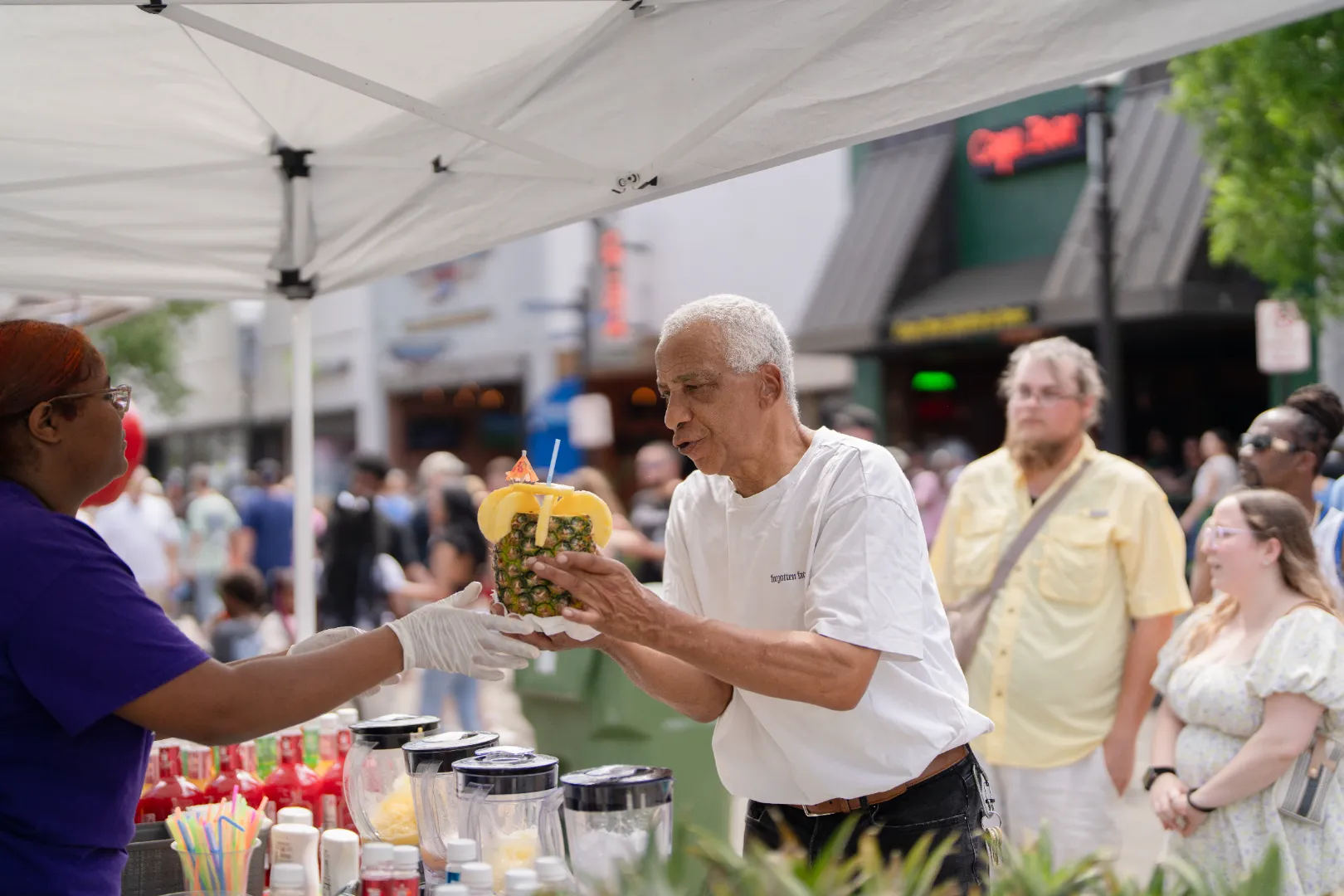 Man in white shirt receiving a decorated pineapple drink from a vendor wearing gloves at an outdoor market.