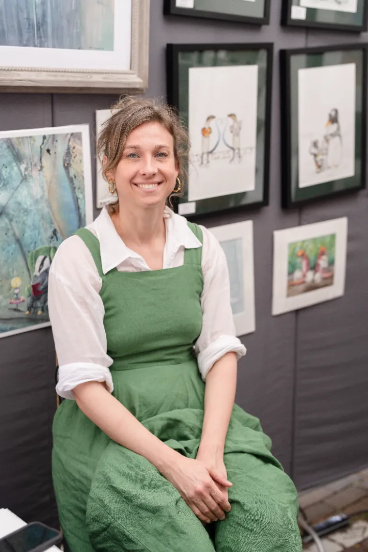 Smiling woman in a green dress and white shirt sitting in front of a wall displaying framed artwork.
