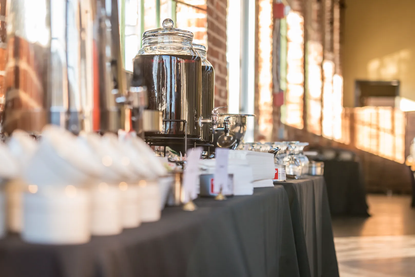 Beverage dispensers and stacked cups arranged on a table covered with a black tablecloth in a sunlit room with brick walls.