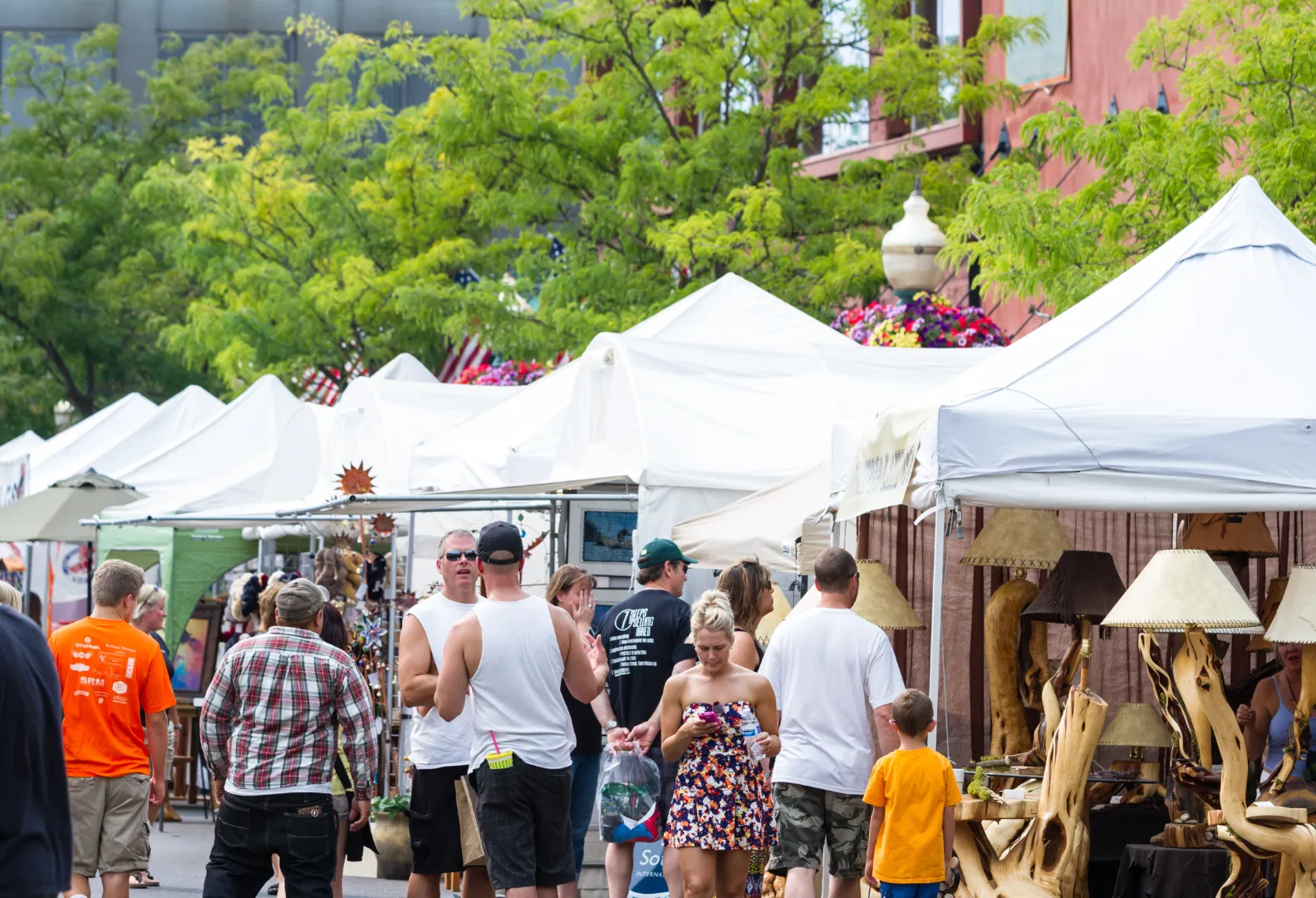 People walking and browsing at an outdoor market with white tents and wooden lamps on display.