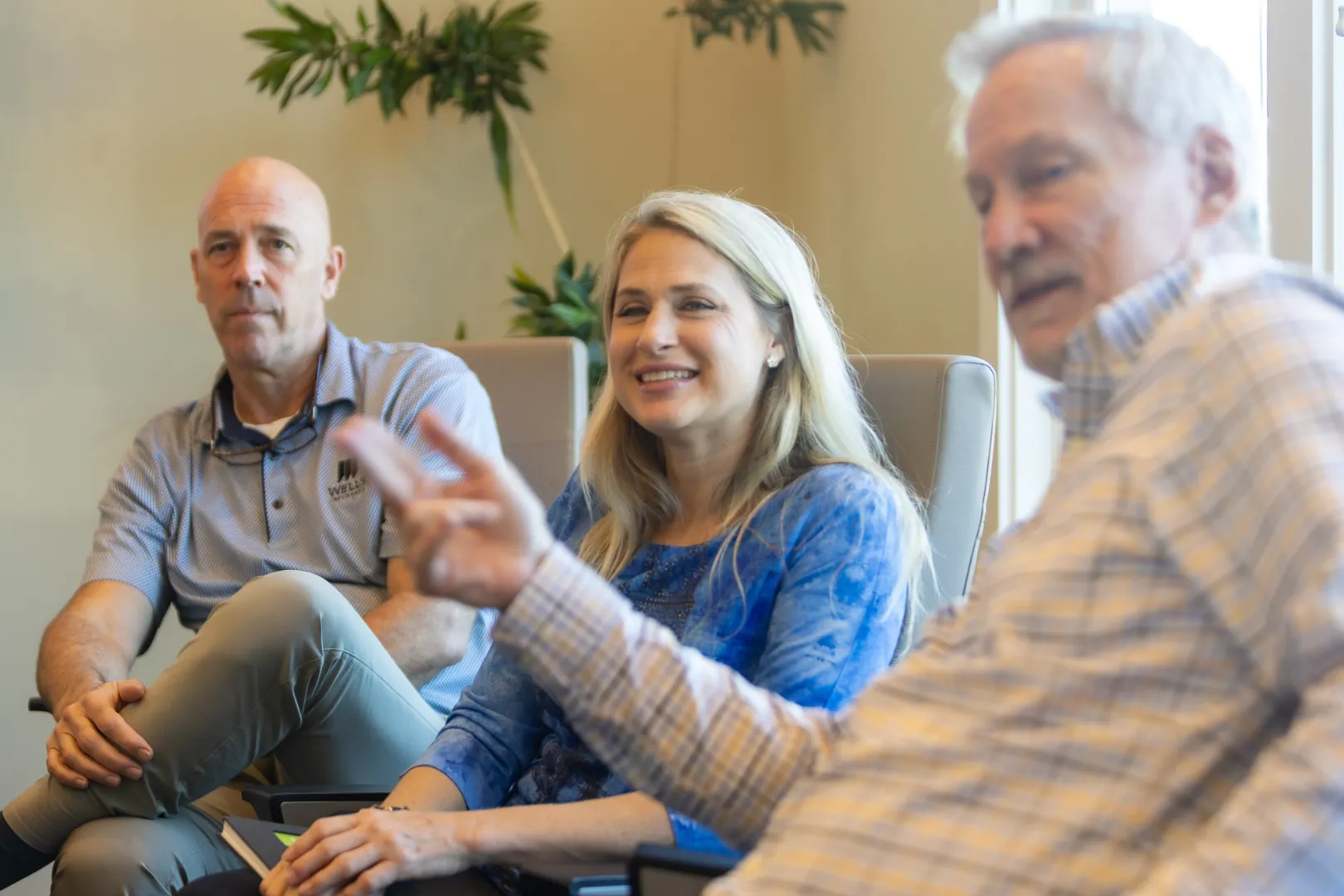 Three people seated indoors, with a smiling woman in a blue shirt in the center and two men on either side.