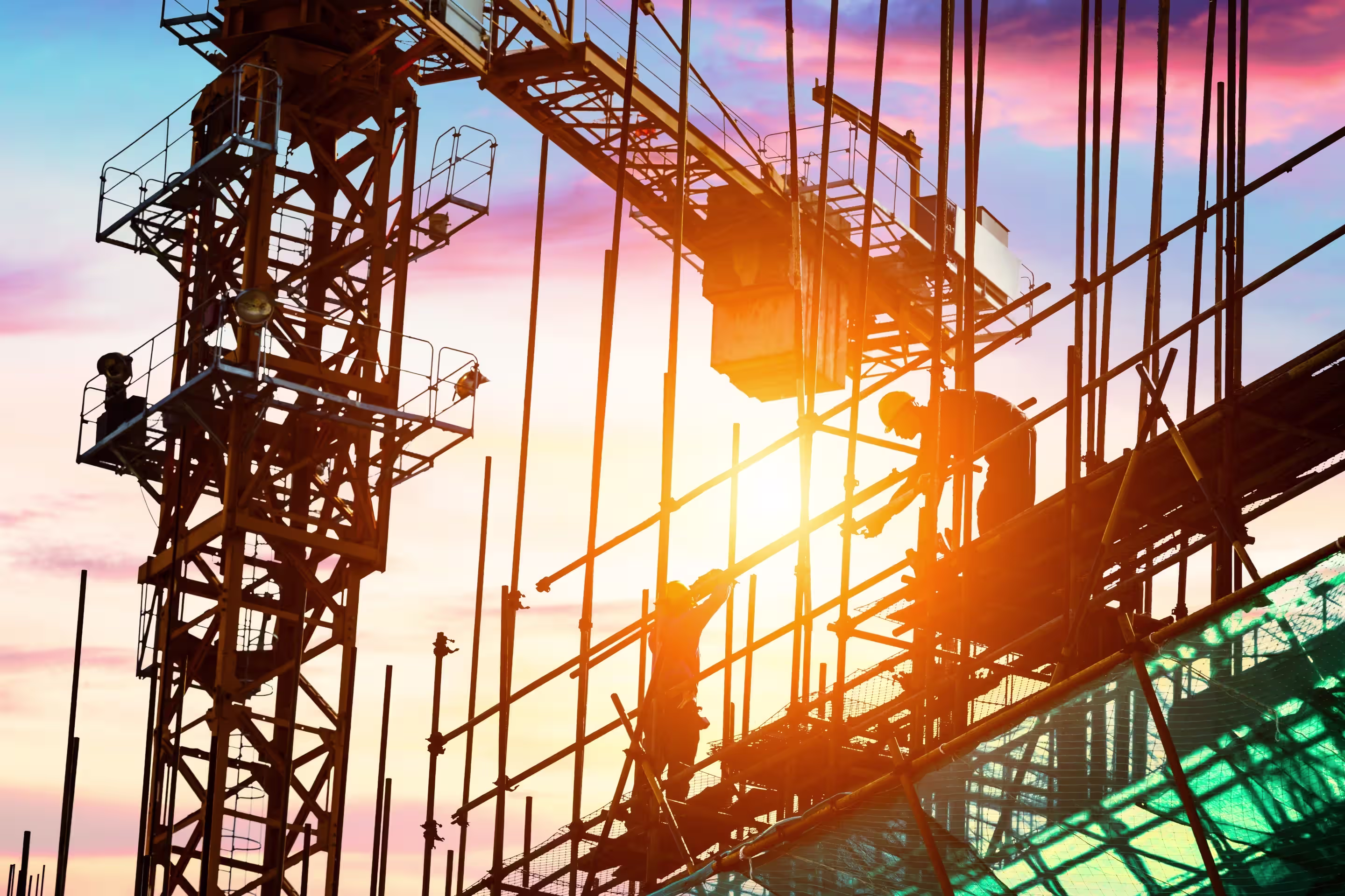 Construction workers adjusting scaffolding at a building site during sunset with a crane in the background.