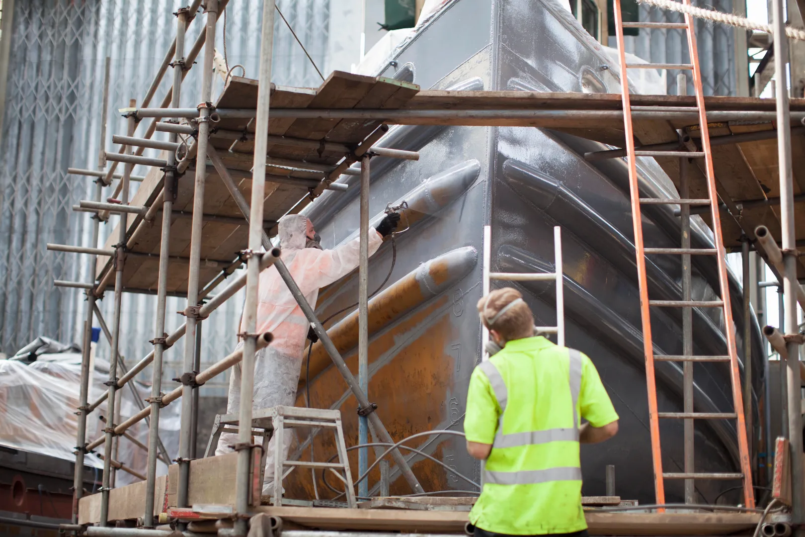 Worker in white protective suit spray-painting the hull of a large ship inside scaffolding, while another worker in a neon yellow safety vest watches.