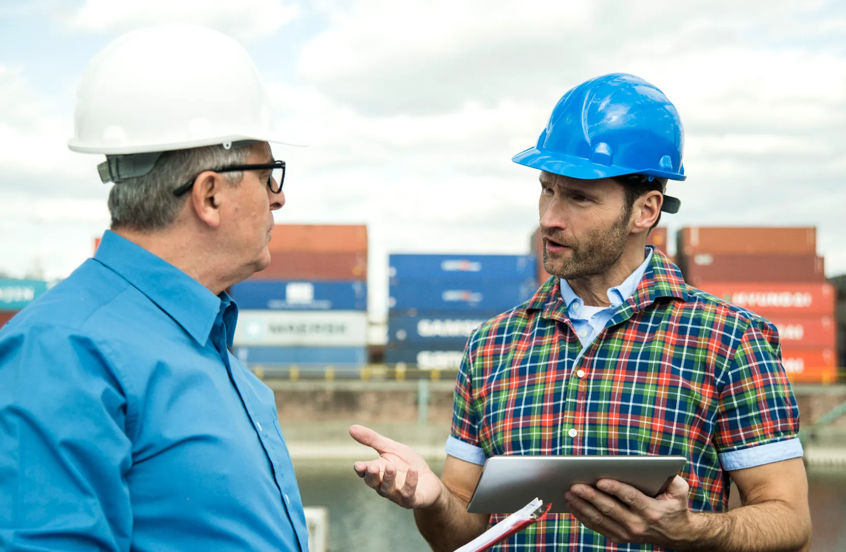 Two male workers wearing safety helmets discussing near stacked shipping containers, one holding a tablet.
