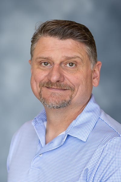 Smiling man with short wavy gray hair and a goatee wearing a blue button-up shirt against a blurred gray background.