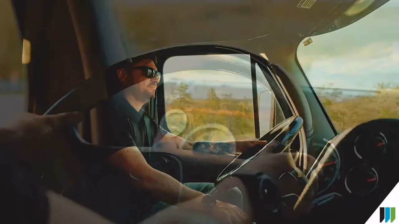 Man wearing sunglasses drives a truck, hands on the steering wheel, dashboard gauges, with a sunny rural landscape outside.