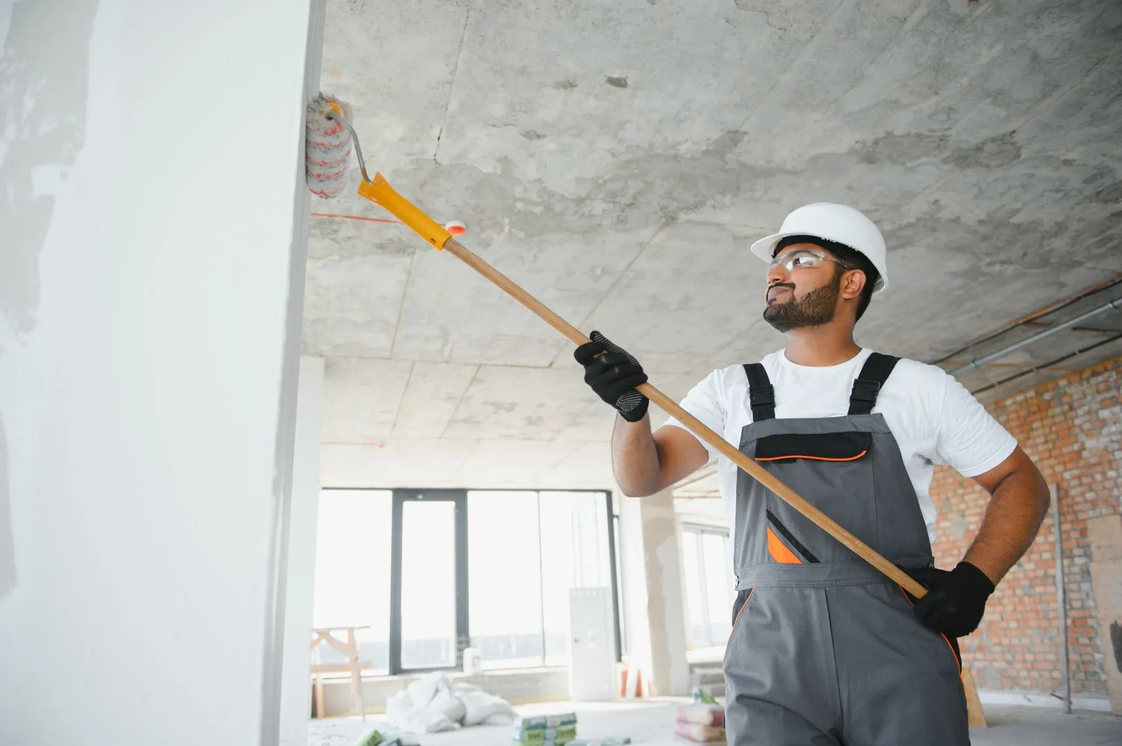 A photograph of a man in a hardhat rolling white paint onto a wall
