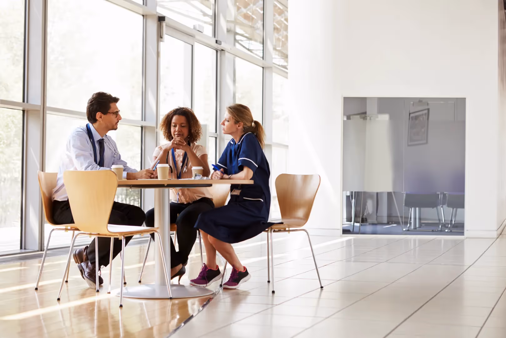 A photograph of three people sitting at an office cafeteria table having a conversation over coffee