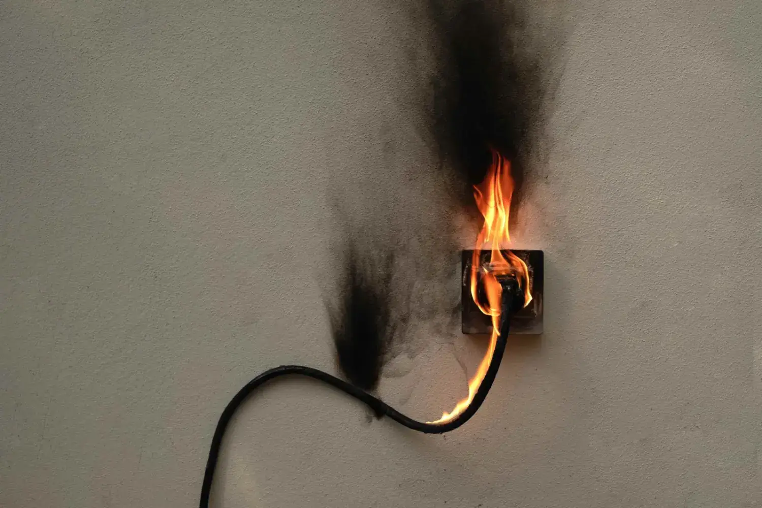 Flames engulf a wall outlet as a thick black cord burns, smoke billowing from a scorched beige wall.